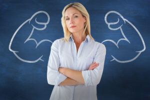 A woman standing with crossed arms in front of a chalkboard with muscular arms drawn on it.