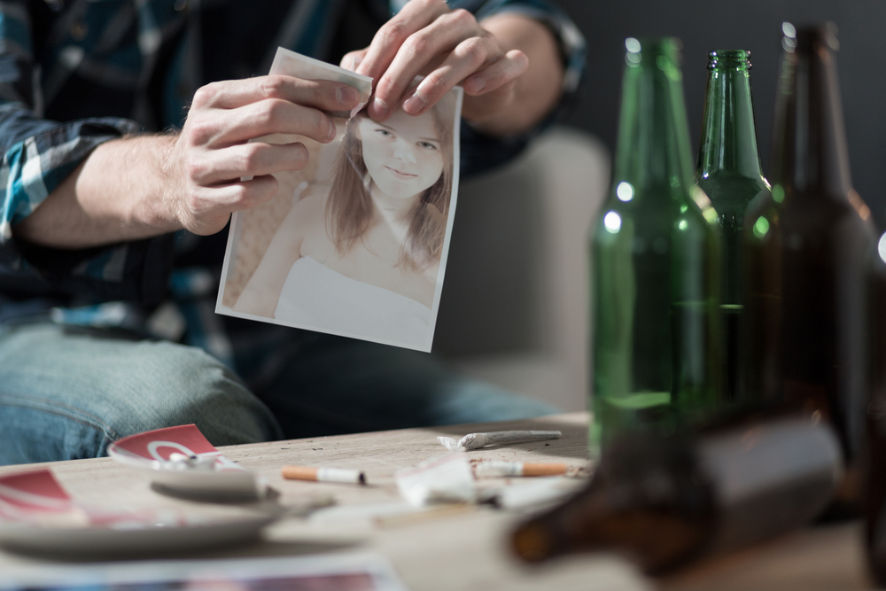 Hands ripping an image of a woman's face and a table with bottles and cigarettes.