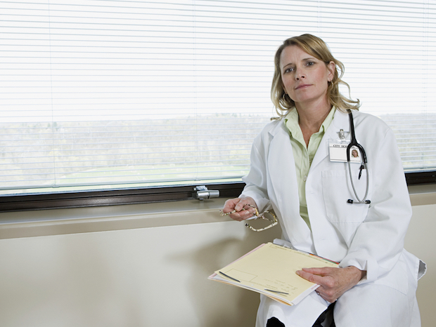 A doctor sitting at a window, holding a folder and glasses.