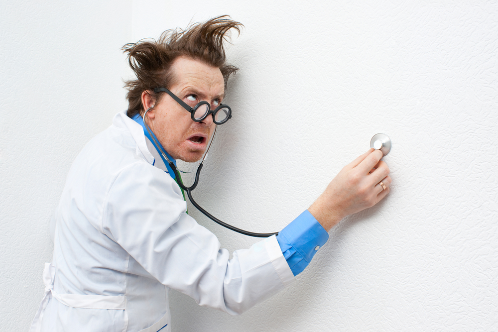 A doctor with wild hair holds his stethoscope against a wall.