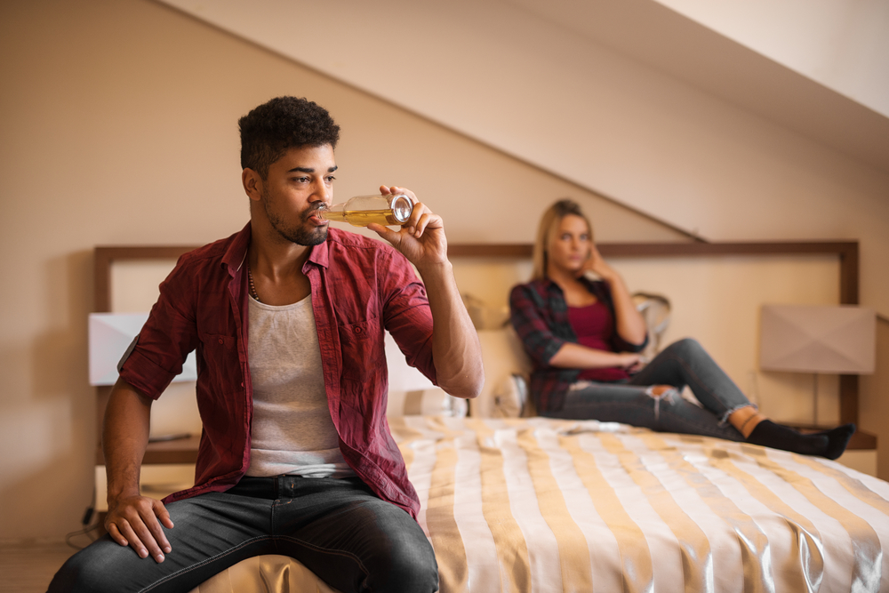 Man drinking on bed while woman in background is angry.