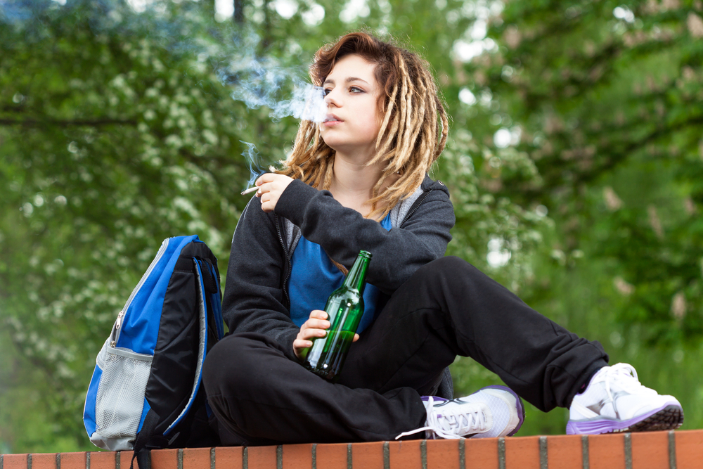 A young person sitting on a wall with a bottle in one hand, cigarette in the other, exhaling smoke.