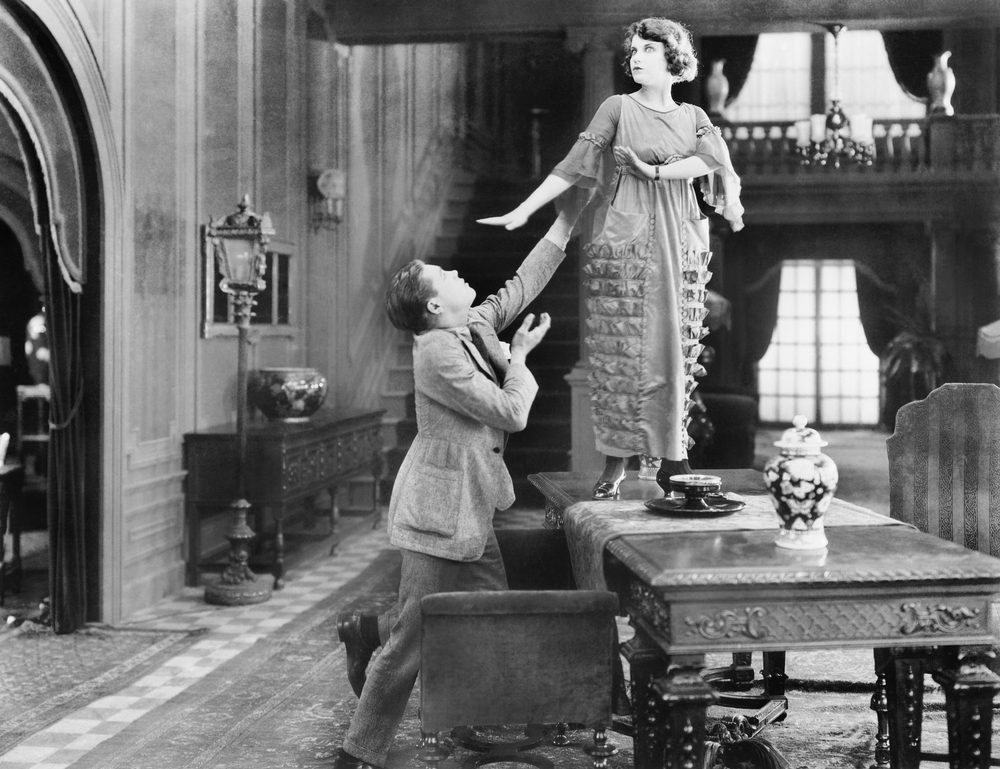 Old time black and white image of a man beseeching a woman who is standing on a table.