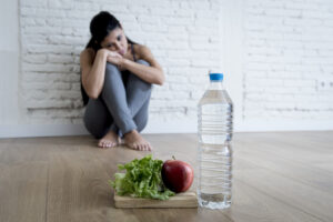 A woman sitting against a wall, looking at a plate of vegetables and a bottle of water several feet in front of her