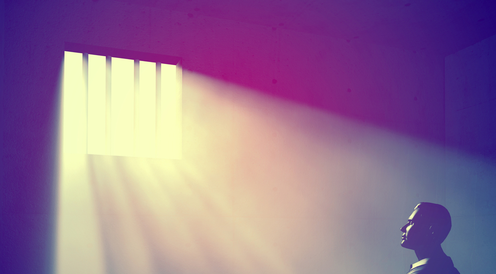 The silhouette of a man staring up through a barred window, light shining in.