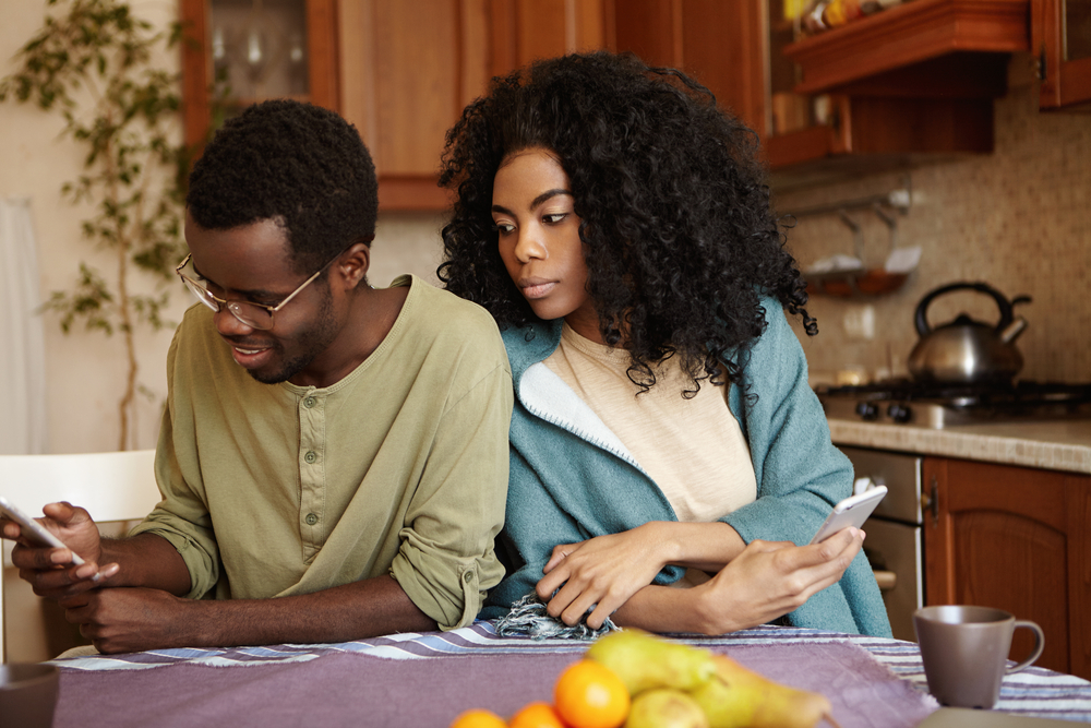 A man looking down at his phone; a woman next to him, looking over his shoulder at the phone, suspicious