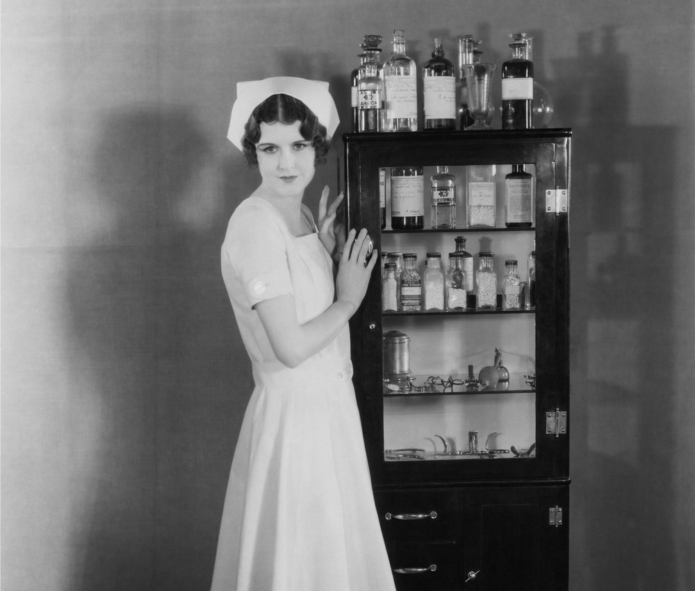 A nurse from the early 20th century standing in front of a medicine cabinet.
