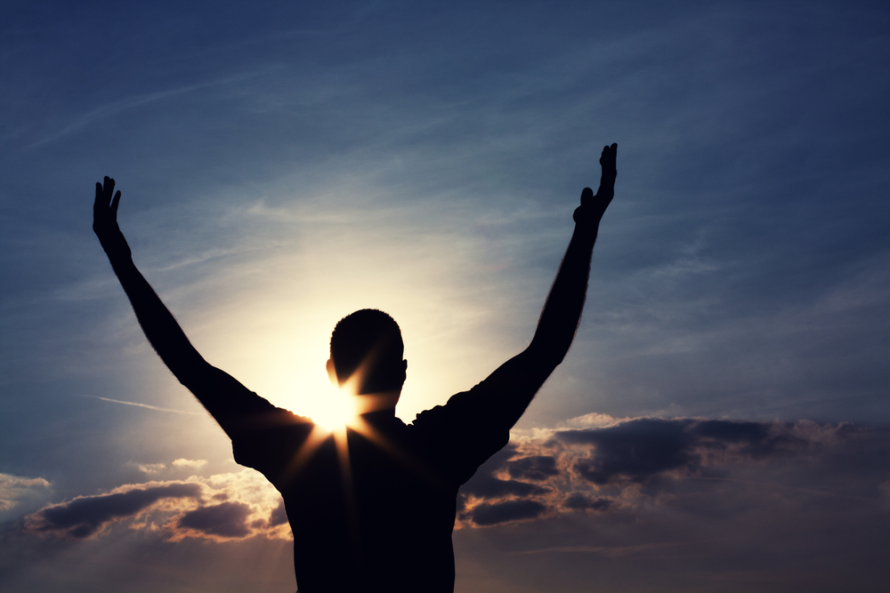 A man with his arms up, the sun behind him.