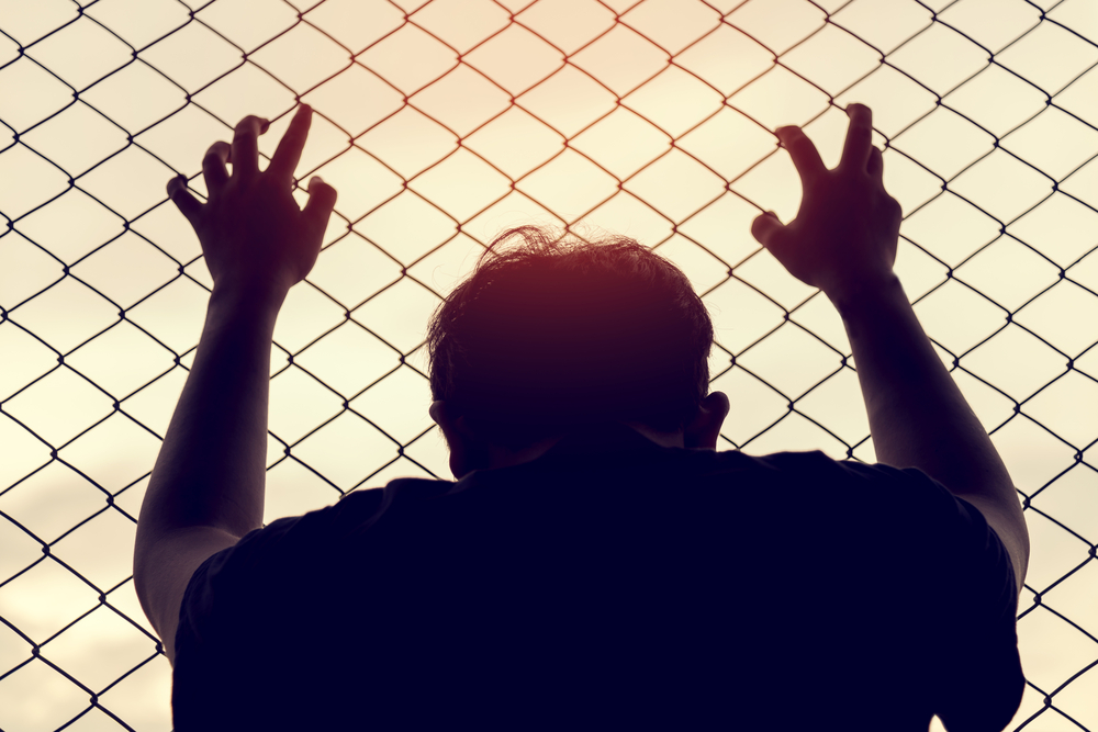 A man in shadow holding onto a chain link fence.