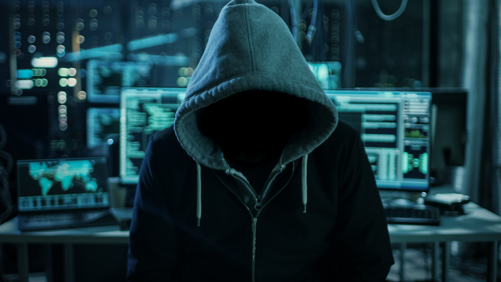 hooded man standing in front of a dark operating room with multiple computer displays