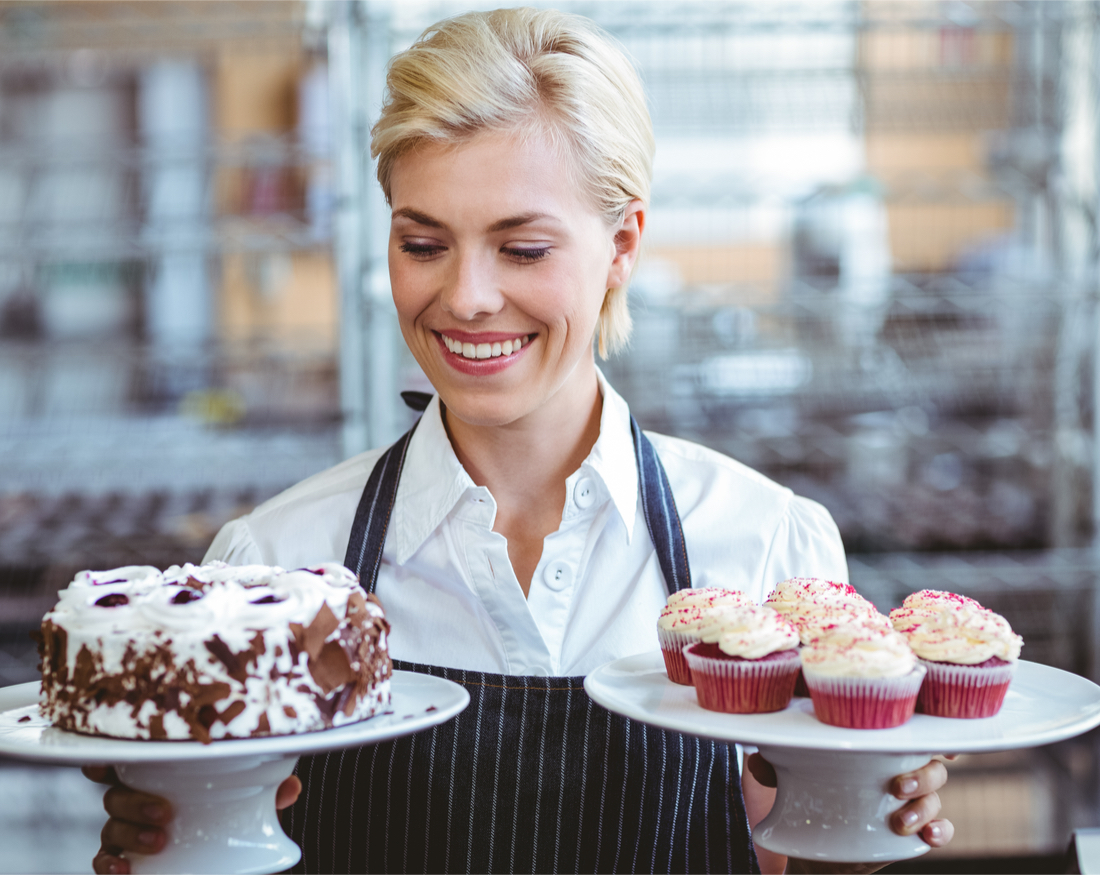 baker holding a platter of cupcakes and a cake platter.