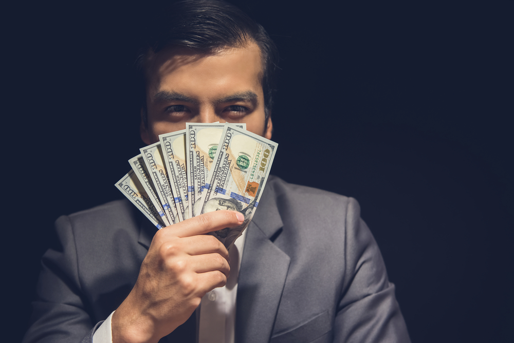 Sly businessman hiding his face behind US dollar banknotes in the dark