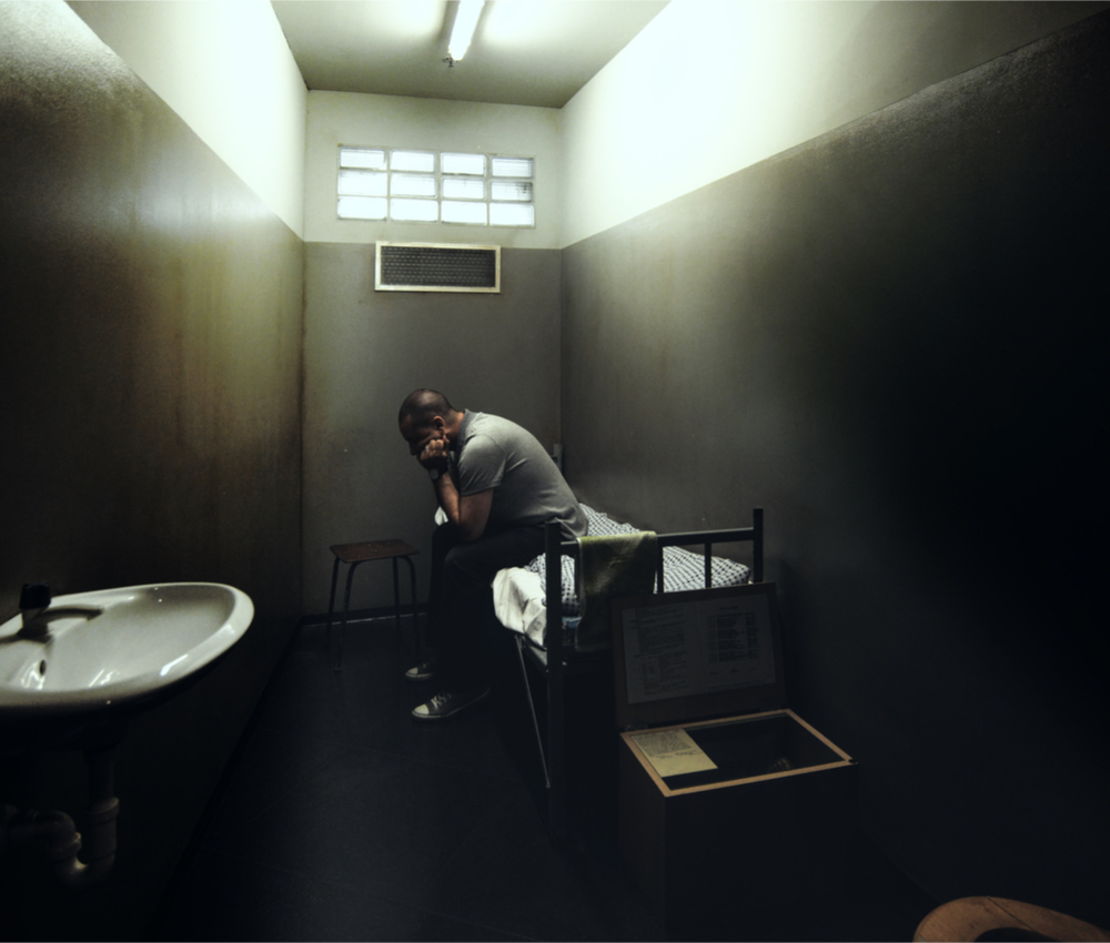 man in prison cell sitting on bed.