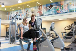 Two women in a gym, one on an exercise bike.