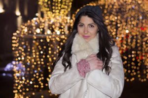 A woman in a white coat, in front of holiday lights.