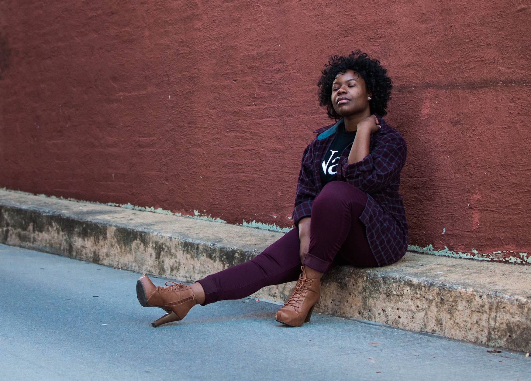 Woman with eyes closed sitting on a curb and against a wall.