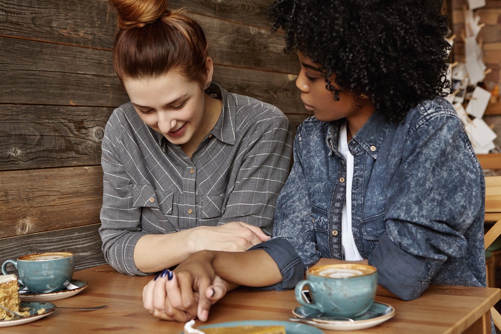 Two women sit at a table, holding hands and drinking coffee