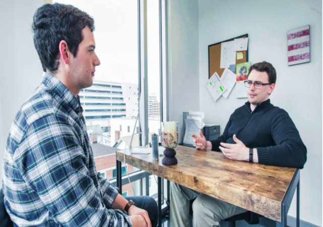 A man at a desk, counseling a younger man.