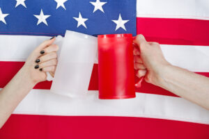 Close up image of both female and male hands holding large plastic drinking mugs with United States of America flag in background