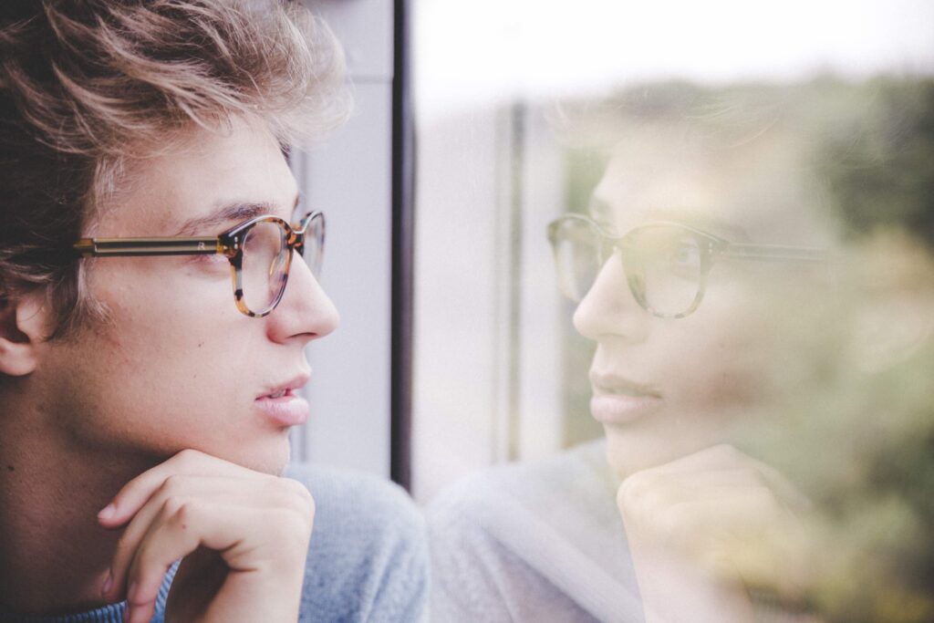 Boy with glasses looking at his reflection in window.