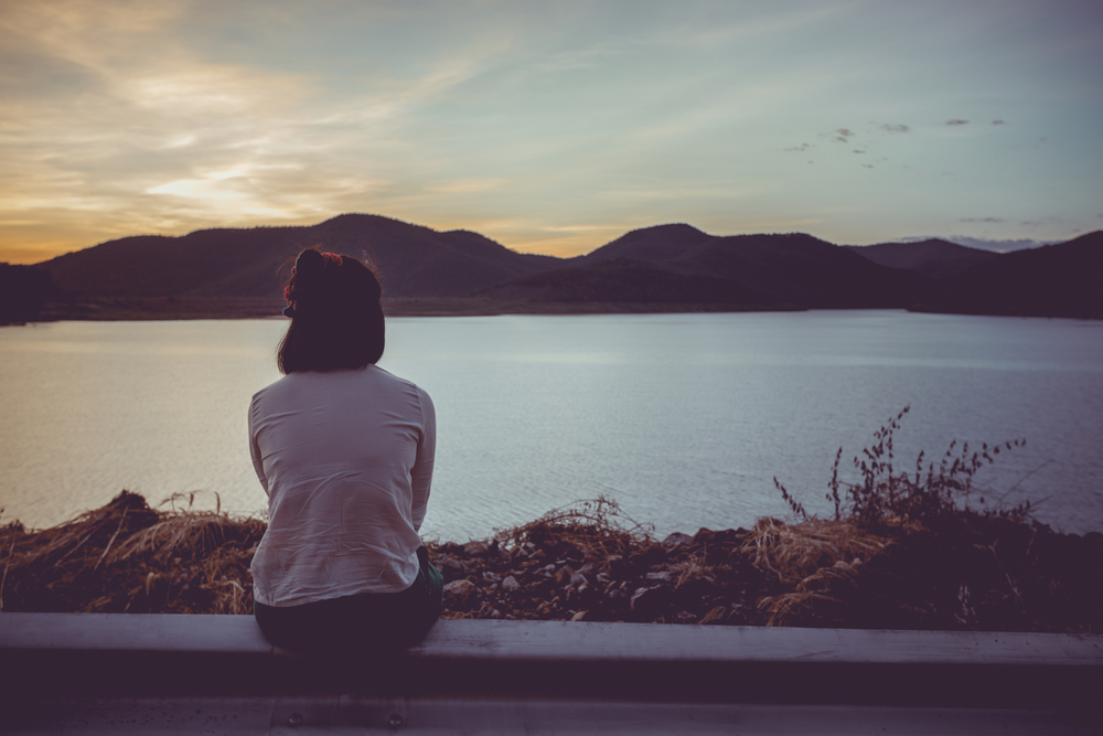 woman seated, facing a body of water and mountains