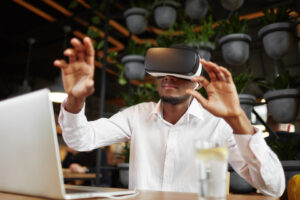 Man wearing virtual reality goggles and gesturing in front of a computer.
