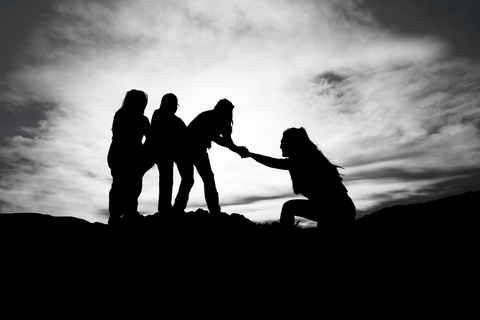 Silhouette of 3 climbers reaching down to help the 4th one up.