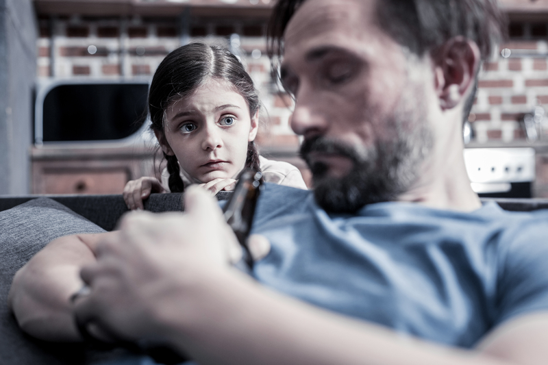 Little girl looks over shoulder of man with eyes closed, holding a beer bottle.