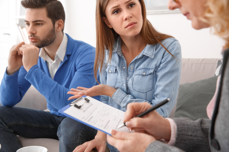 Man and woman sit on couch, therapist sits on chair with clipboard.