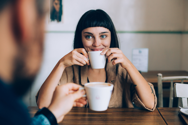 Woman smiles over coffee cup at man across the table.