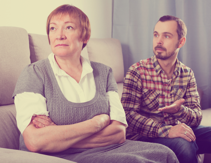 Older mother sits with adult son on couch. They look like they're mid-argument.