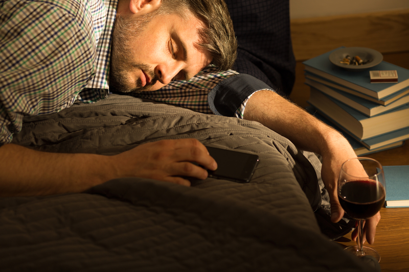 Man is passed out on bed with glass of wine. A stack of books is next to the bed.