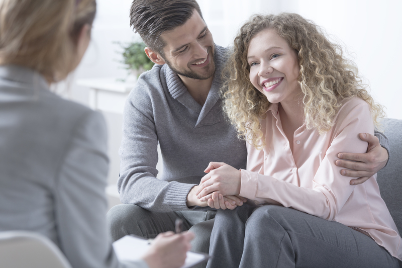 A smiling couple sits on a couch opposite a therapist.