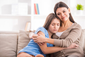 Mother and sad teen daughter hugging on sofa at home.