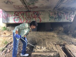 A woman examines the ground with a tool under a graffiti-covered bridge