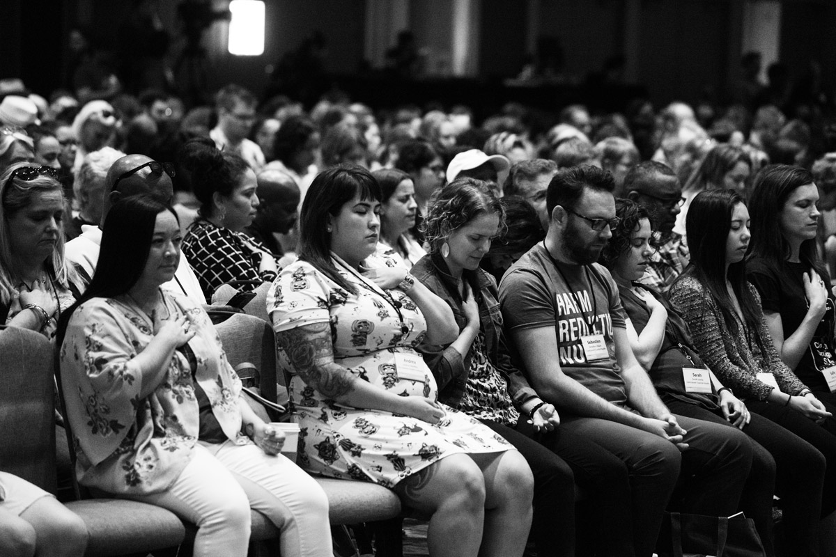 Audience members in an auditorium sit with hands over hearts, some with eyes closed.
