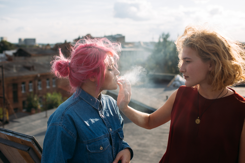 2 girls, one is holding the cigarette in the other's mouth.