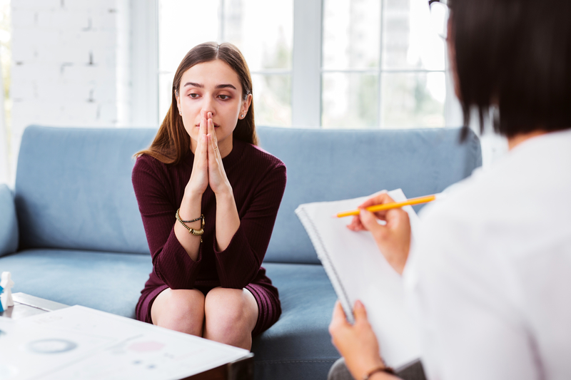 Worried woman sits on couch, facing professional or doctor.