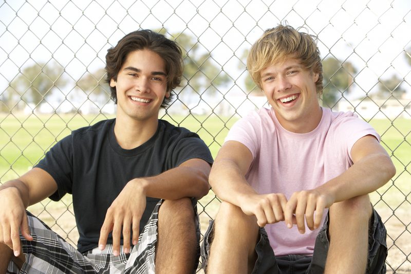 2 teen boys sitting against a fence, smiling.
