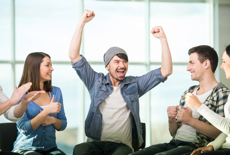 Group of people in circle, one person has arms up with joy, others clap, 12-step meeting or AA