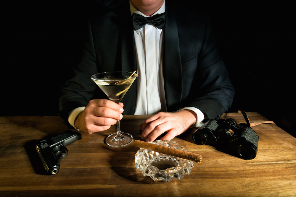 James Bond sitting in front of table with alcohol, gun, and cigars.