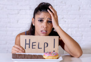 Woman with sugar addiction, has upset face and "Help!" sign in front of plate with sweets.