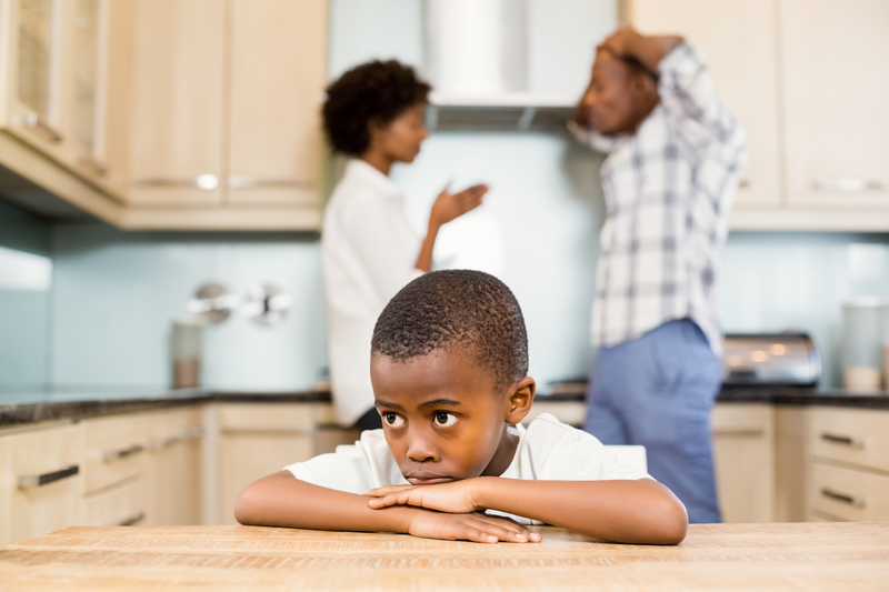 Child sitting, chin on hands, while parents gesture in background. Family with mental illness