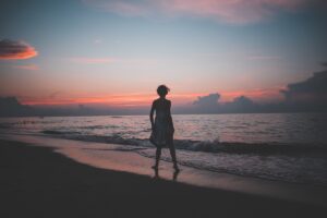 Woman at shoreline in front of setting sun, clouds, ocean