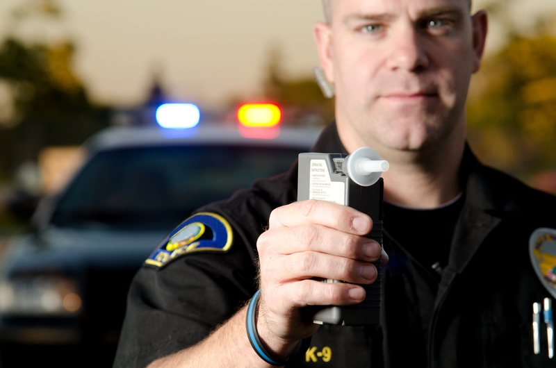 Police officer stands in front of patrol car while holding up breathalyzer