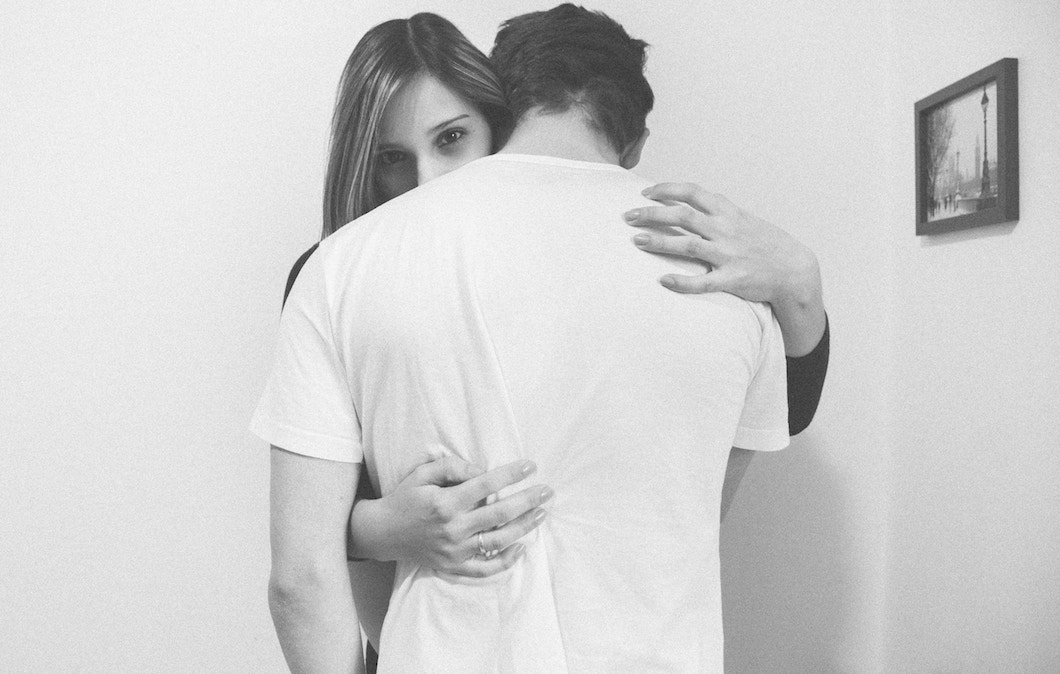 Black and white image of couple embracing, woman's face visible, facing camera, over man's shoulder.