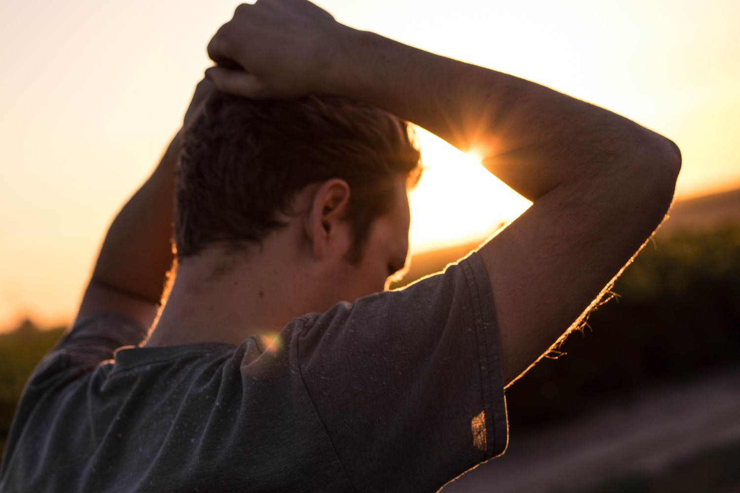 Man with hands on head, looking ahead, silhouetted by setting sun