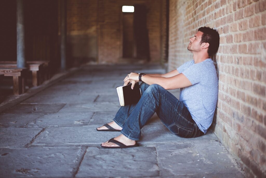 The 5 Most Common Myths About Faith-Based Addiction Treatment Programs Man sitting against brick wall, book or bible in hand