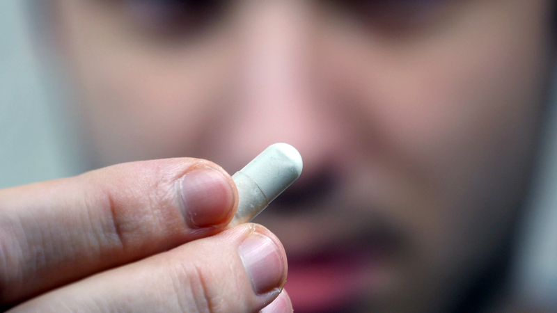 Close up of hand holding pill in front of man's face.