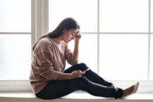 Woman looks sad or depressed, sitting at window looking at smartphone.